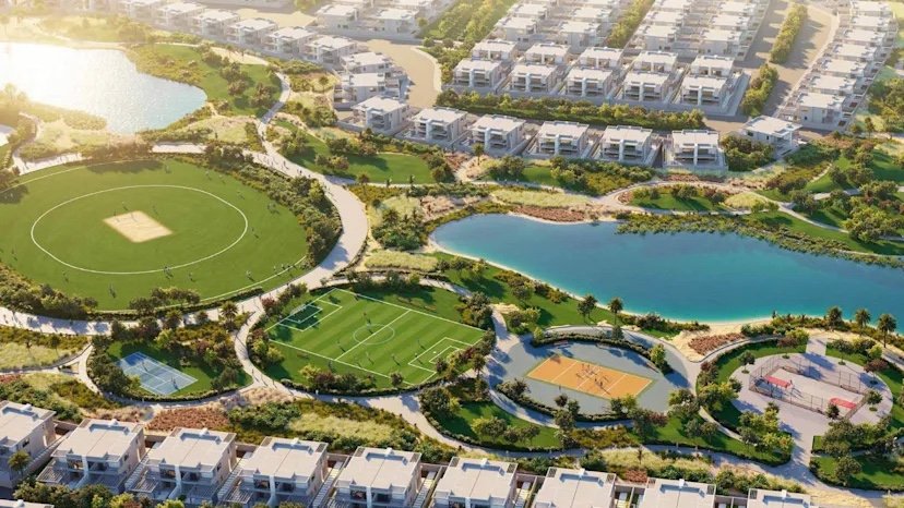 Aerial view of Damac Hills 2 townhouses around a lake, with a cricket ground, football pitch and sports courts